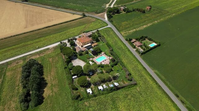 An aerial drone view reveals a charming Tuscan agriturismo nestled among vibrant sunflower fields and vast farmland. The property features a swimming pool and an organized camperstop for motorhomes in