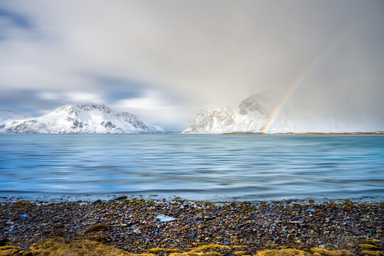 View of the Lofoten islands featuring snow-covered mountains, a rainbow over the calm sea, and a rocky shoreline in Nordland, Norway.