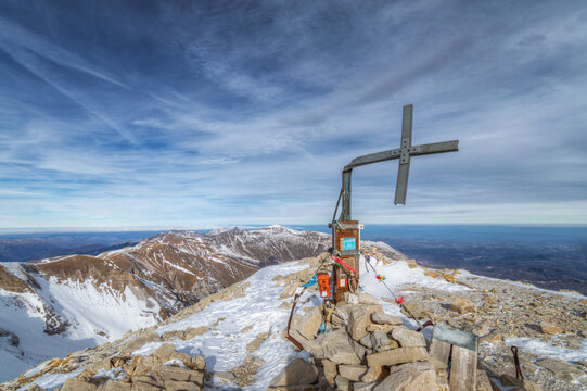 View of the summit cross surrounded by snow-covered peaks and a vast mountain landscape under a blue sky in Monte Vettore, Marche, Italy.