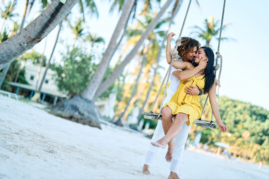 A happy couple embraces on a wooden swing at a tropical beach, sharing a tender romantic moment. Palm trees, white sand and warm sunlight create a relaxed summer vacation mood as they laugh and hold e