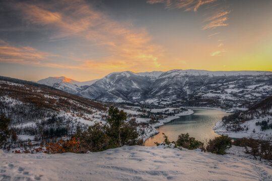 View of the snow-covered mountains and the calm waters of Lago di Fiastra during a golden sunset in Fiastra, Marche, Italy.