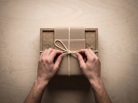 Top view of male hands tying a bow with twine string around a wooden gift box wrapped in eco-friendly corrugated craft paper
