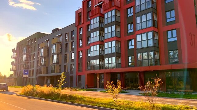 Modern apartment building block with red and grey facade. Sunlight illuminates geometric architecture and young trees along paved walkway. Contemporary housing development exterior design. 