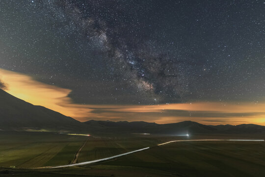 View of the Milky Way galaxy stretching over the dark mountains and the vast plains with light trails from a car on the road in Castelluccio, Umbria, Italy.