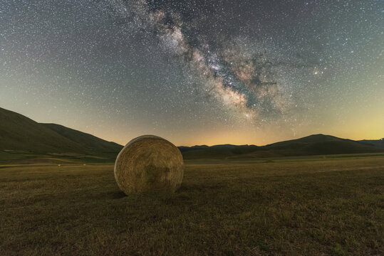 View of a solitary hay bale in the vast fields of the Piano Grande under a brilliant starry sky with the Milky Way galaxy visible above the rolling hills of Castelluccio, Umbria, Italy.