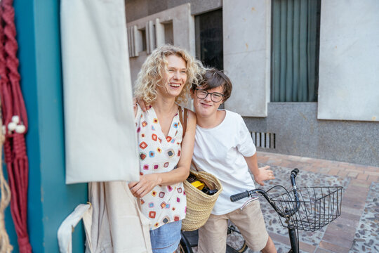 Joyful mother and teenage son with Down syndrome sharing playful moment beside bicycle in Spain