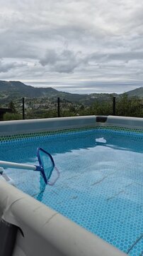 ​Cleaning blue water in an above-ground swimming pool using a skimmer net with beautiful mountain view in the background during summer pool maintenance.