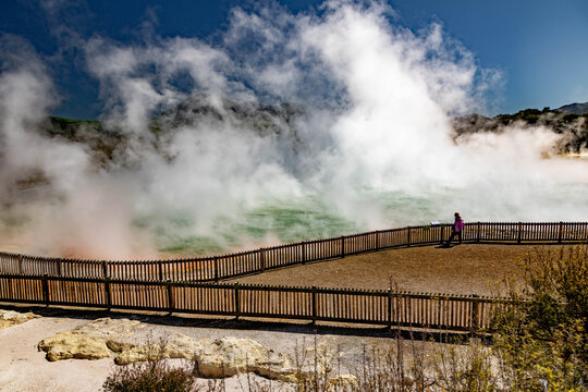 View of the geothermal pool at Waiotapu Thermal Wonderland with rising steam, a wooden fence, and a person standing nearby in Rotorua, Waikato Region, New Zealand.