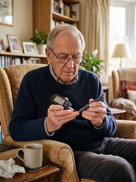 Elderly man in armchair holding glucometer and lancing device to perform blood sugar medical test at home. Insulin dependent diabetes, medical symptoms and lifestyle diabetic concept, copy space.