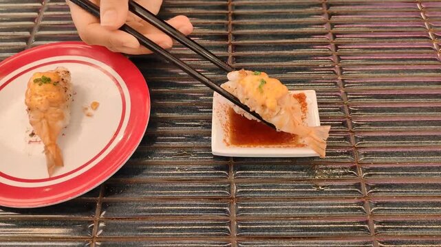 Hand using chopsticks to dip shrimp nigiri sushi into sauce at a Japanese restaurant.