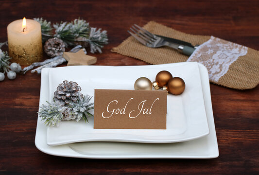 Elegant Scandinavian Christmas table setting with burning candle, rustic burlap napkin, gold baubles and place card with text God Jul on a white plate.