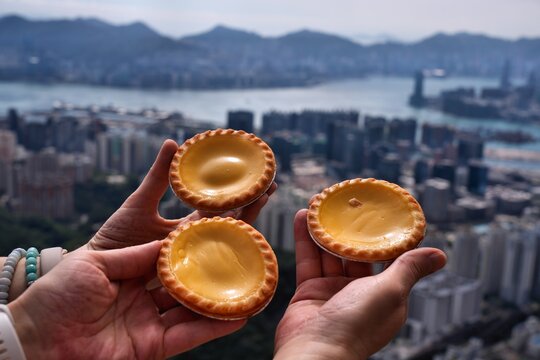 Hands holding Hong Kong egg tarts. Kowloon and Hong Kong cityscape with Victoria Harbour in background.