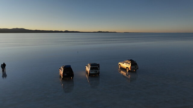 Potosi Department, Bolivia - 01 October 2026: Aerial view of Uyuni Salt Flat with three SUVs parked on the reflective surface and people standing nearby under the golden hour light.