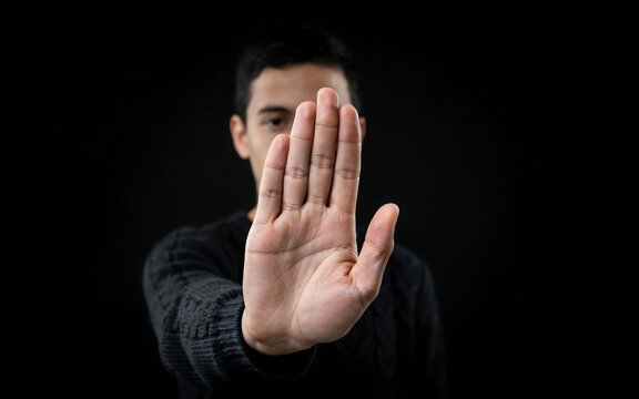 Young man making stop gesture with his open palm facing forward in dark studio environment