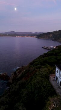 Aerial view of Getariako Itsasargia lighthouse on the rocky coastline of Getaria at dusk with the moon visible in the sky Getaria, Euskadi, Spain.