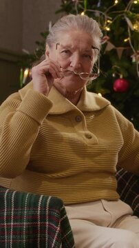 Portrait of senior Caucasian woman putting on eyeglasses near Christmas tree. Footage suitable for holiday vision care advertisements and senior healthcare campaigns, vertical