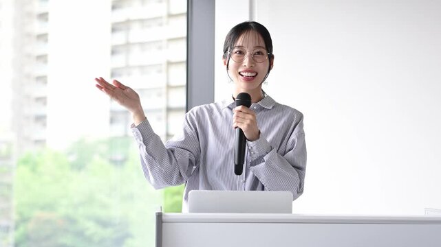会議室でマイクを持ってプレゼンテーションを行う30代の日本人ビジネスパーソンの女性