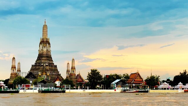 Wat Arun temple rises beside the Chao Phraya River in Bangkok, Thailand, as a long-tail boat cruises past during a peaceful evening with soft clouds and warm sunset light over the riverside.