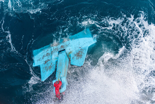 Heavy commercial ship anchor emerging from churning blue seawater, with powerful anchor chain links lifting the massive metal structure above the surface.
