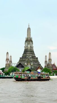 Wat Arun temple rises beside the Chao Phraya River in Bangkok, Thailand, as a long-tail boat cruises past during a peaceful evening with soft clouds and warm sunset light over the riverside.