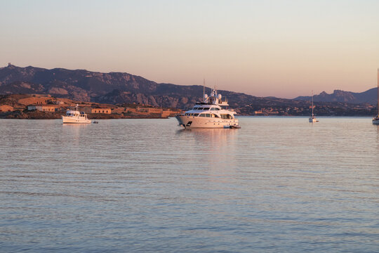 View of luxury yachts anchored in the calm waters of Cala di Punta Rossa at sunset, with rolling hills and coastal landscape in the background in La Maddalena, Sardegna, Italy.