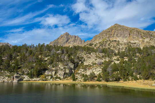 View of Llac de Pessons with rocky mountain peaks, pine trees, and calm water reflections in the Pyrenees in Grau Roig, Encamp, Andorra.