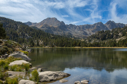 View of Llac de Pessons surrounded by pine forests and rugged mountain peaks under a blue sky with wispy clouds in Grau Roig, Encamp, Andorra.