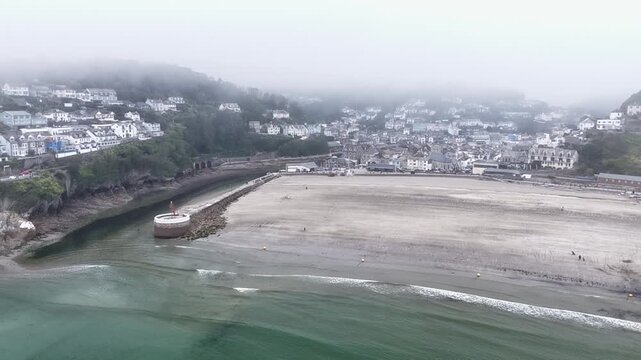 Aerial view of Looe beach on a foggy day with the historic town and harbour visible under a thick mist in Looe, England, United Kingdom.