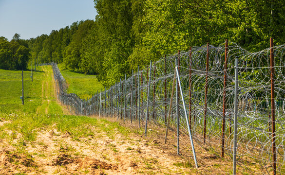 View of a border fence with razor wire extending through a grassy field and along the edge of a forest in Europe.