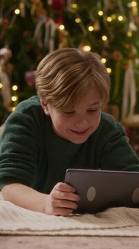 Vertical video of Caucasian boy lying on floor, watching digital tablet near illuminated Christmas tree. Footage suits mobile app advertising, holiday gaming promos, or digital education campaigns