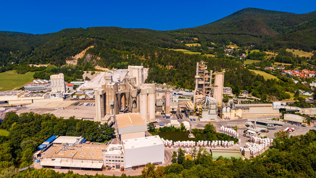 Processing towers stand at cement works below wooded hills.. Pale warehouses and gray silos fill the foreground, with houses and green slopes behind.
