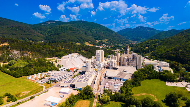 Transport infrastructure runs between buildings at a cement plant.. A straight transport corridor runs through pale factory buildings beside green fields and wooded slopes.