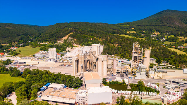 Silos rise beside a steel tower at an Austrian cement plant.. Gray cylindrical structures and open steel framing rise above low roofs beside green woodland.