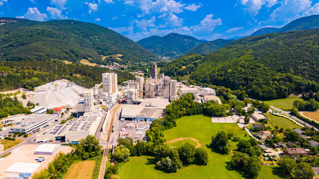 A cement factory borders a meadow below mountain slopes.. Long pale roofs lead toward gray silos, with trees and green grass beside the industrial site.