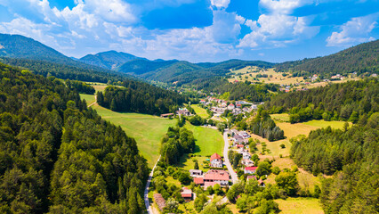 Obraz premium A village road crosses farmland in a wooded Austrian valley.. Red roofs line a curving road among green and yellow fields beneath scattered cloud shadows.