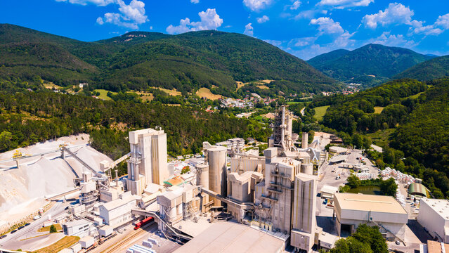Cement silo towers rise below a mountain ridge.. Gray processing structures rise above tan roofs, with green hills under a blue sky.