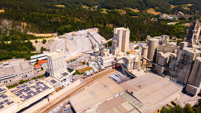 Solar panels cover a roof beside cement silos and railway tracks.. Dark panels cover a factory roof beside pale material piles, gray towers and railway tracks.