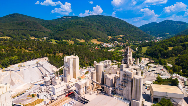 A cluster of cement factory towers rises below mountain slopes.. Pale industrial buildings and gray silos stand below green hills and sunlit white clouds.