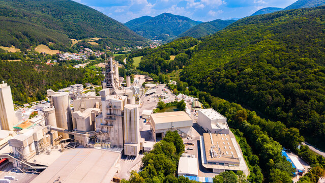 Cement processing towers stand among roofs in a wooded valley.. Gray silos rise beside a broad tan roof, with green hills receding toward distant mountains.