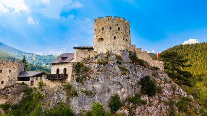 A crenellated Austrian castle keep stands on a rocky outcrop.. Tan masonry and dark roof tiles rise above gray rock, with green woodland and mountains behind. © Vadim