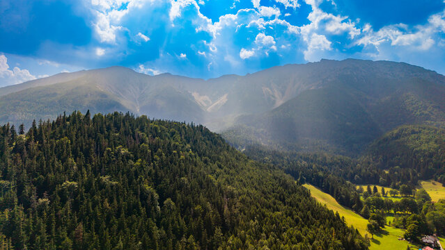 A mountain escarpment rises above a conifer ridge in hazy sunlight.. Pale rock faces rise behind dark green treetops under a blue sky with scattered white clouds.
