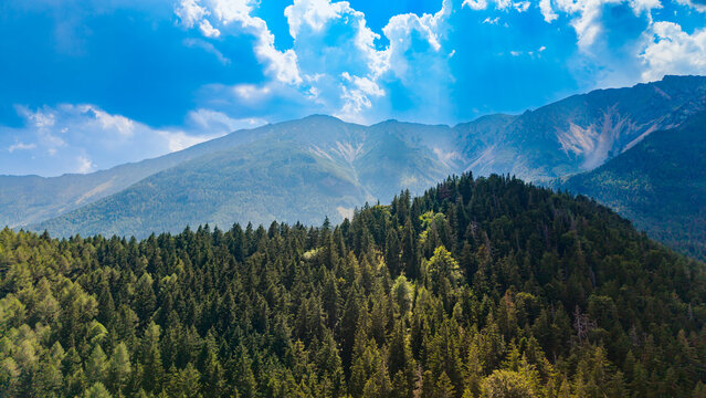 A wooded ridge rises below mountain slopes and scattered clouds.. Green conifers cover a foreground hill below hazy peaks and exposed gray rock channels.