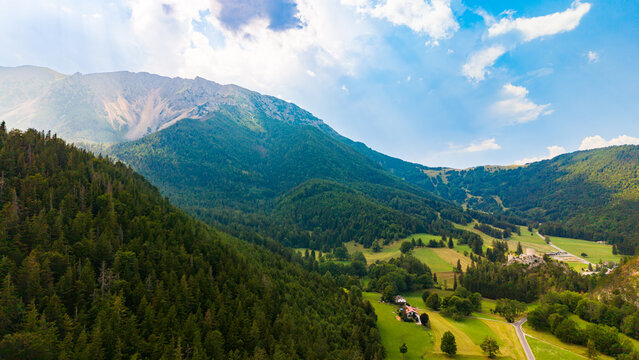 A castle borders farmland between wooded hills in a mountain valley.. A stone tower borders green and yellow fields below hazy ridges and a partly cloudy sky.
