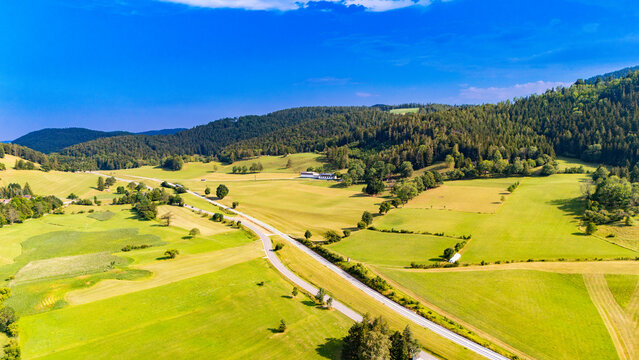 A road runs diagonally through a patchwork of valley fields.. A paved road separates green and yellow plots below wooded hills under a blue sky.