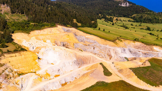 Haul tracks cross rock benches at a terraced Austrian quarry.. Yellow and white rock benches step across an open pit bordered by green fields and woodland.