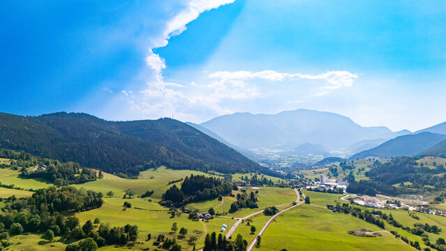 Roads connect industrial buildings among fields in a mountain valley.. Green fields surround pale roofs and curving roads, with hazy ridges beneath scattered clouds.