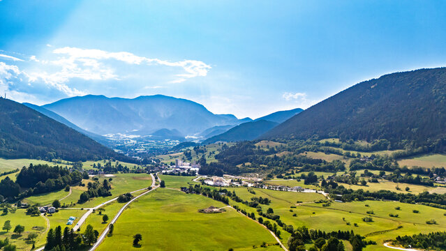 A settlement lies among fields between mountain slopes.. Pale buildings and lanes spread across green farmland beneath layered blue hills and white clouds.