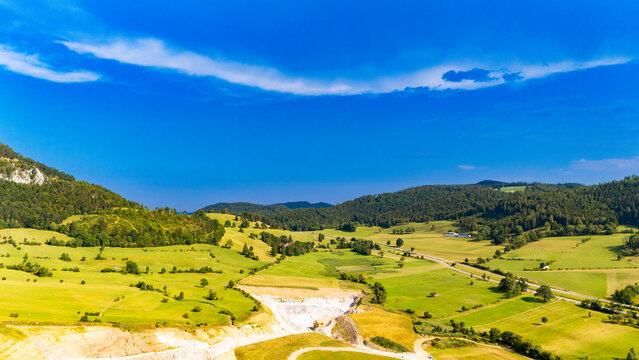 Farmland surrounds a quarry beneath an open sky.. Green meadows surround a pale excavation below wooded hills and a blue sky with thin clouds.