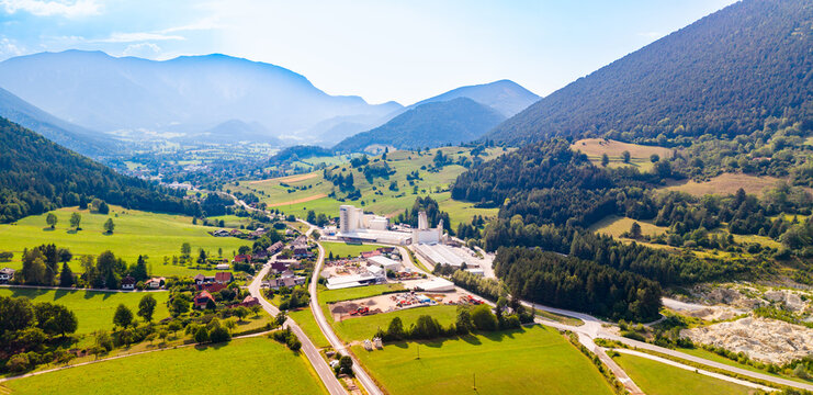 An industrial site stands beside a road in a mountain valley.. Gray towers and pale roofs stand beside a road among green fields beneath hazy ridges.