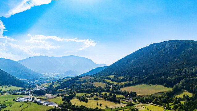 Industrial roofs stand among agricultural fields in a mountain valley.. Pale buildings occupy green farmland below a dark wooded slope and hazy ridges.